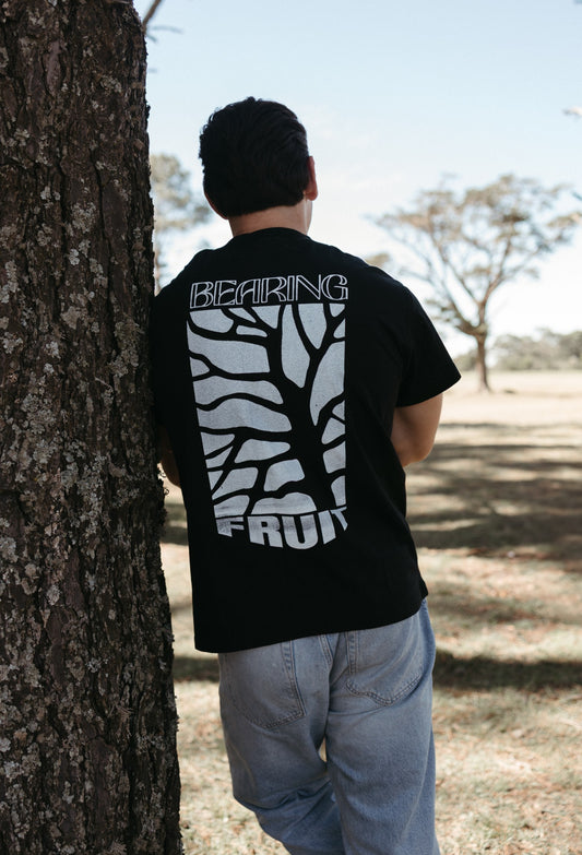 Person wearing a black Bearing Fruit t-shirt with white graphics and text, standing against a tree outdoors.