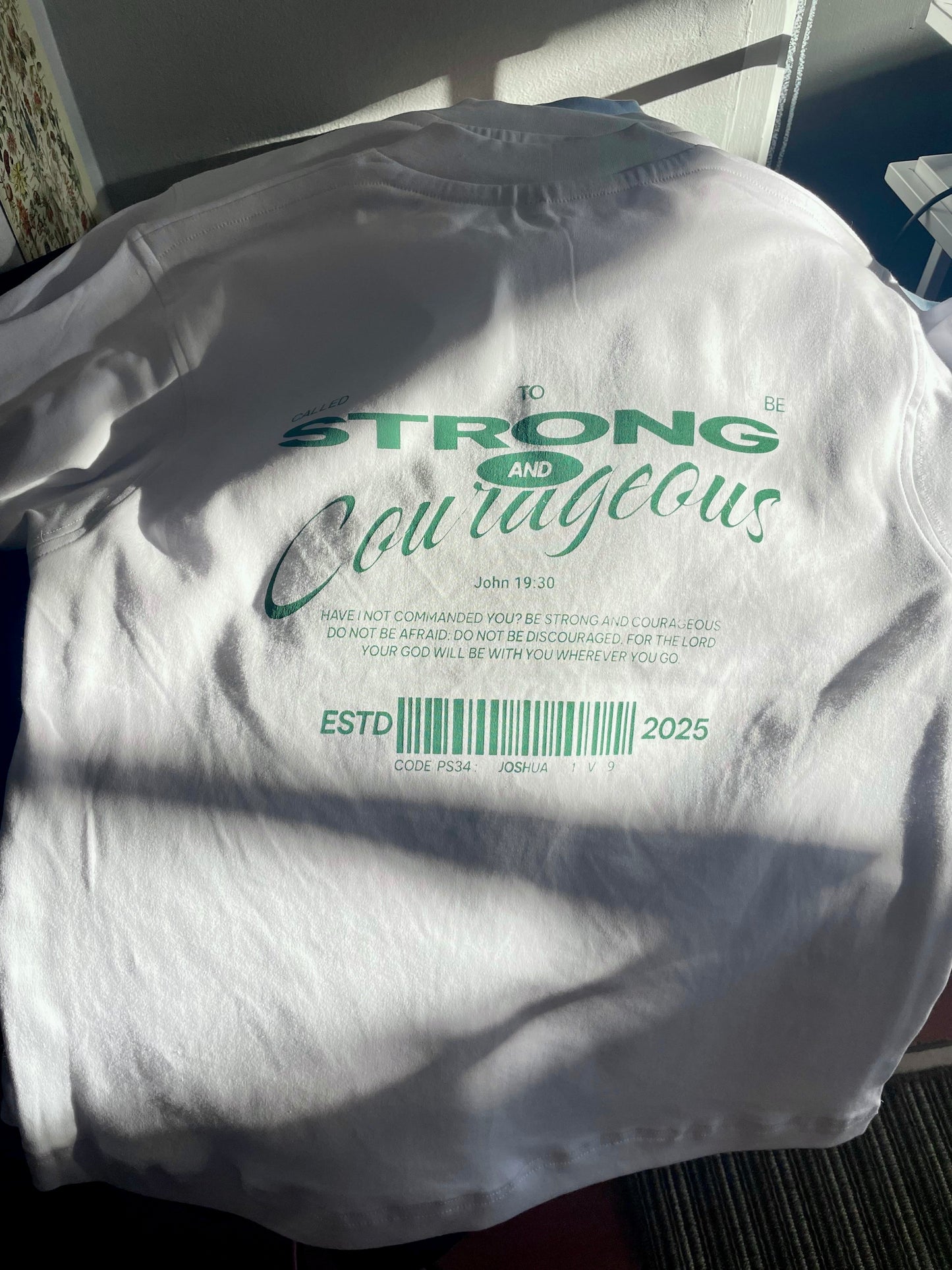 White shirt with text 'STRONG AND COURAGEOUS' and a barcode on a plain background in Port Elizabeth, Eastern Cape, South Africa