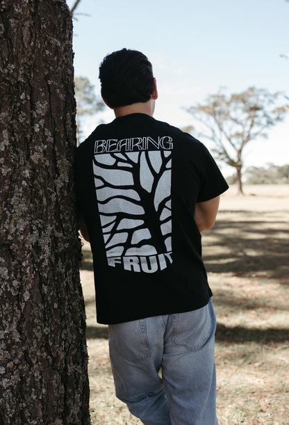 Person wearing a black Bearing Fruit t-shirt with white graphics and text, standing against a tree outdoors.