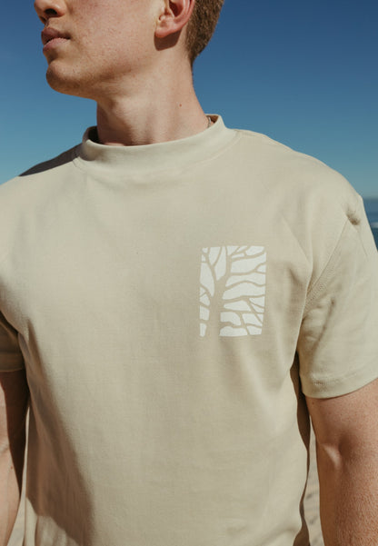 Person wearing a beige Bearing Fruit t-shirt with a white tree design against a blue sky.