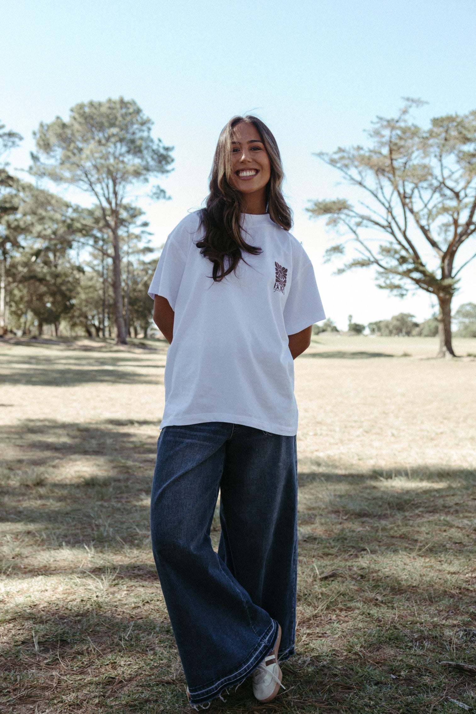 Woman wearing a white Born again oversized T-shirt and blue jeans standing in a field with trees in the background