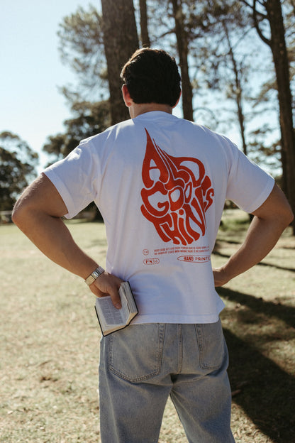Person wearing a white Christian All Good Things t-shirt with orange graphics and text outdoors