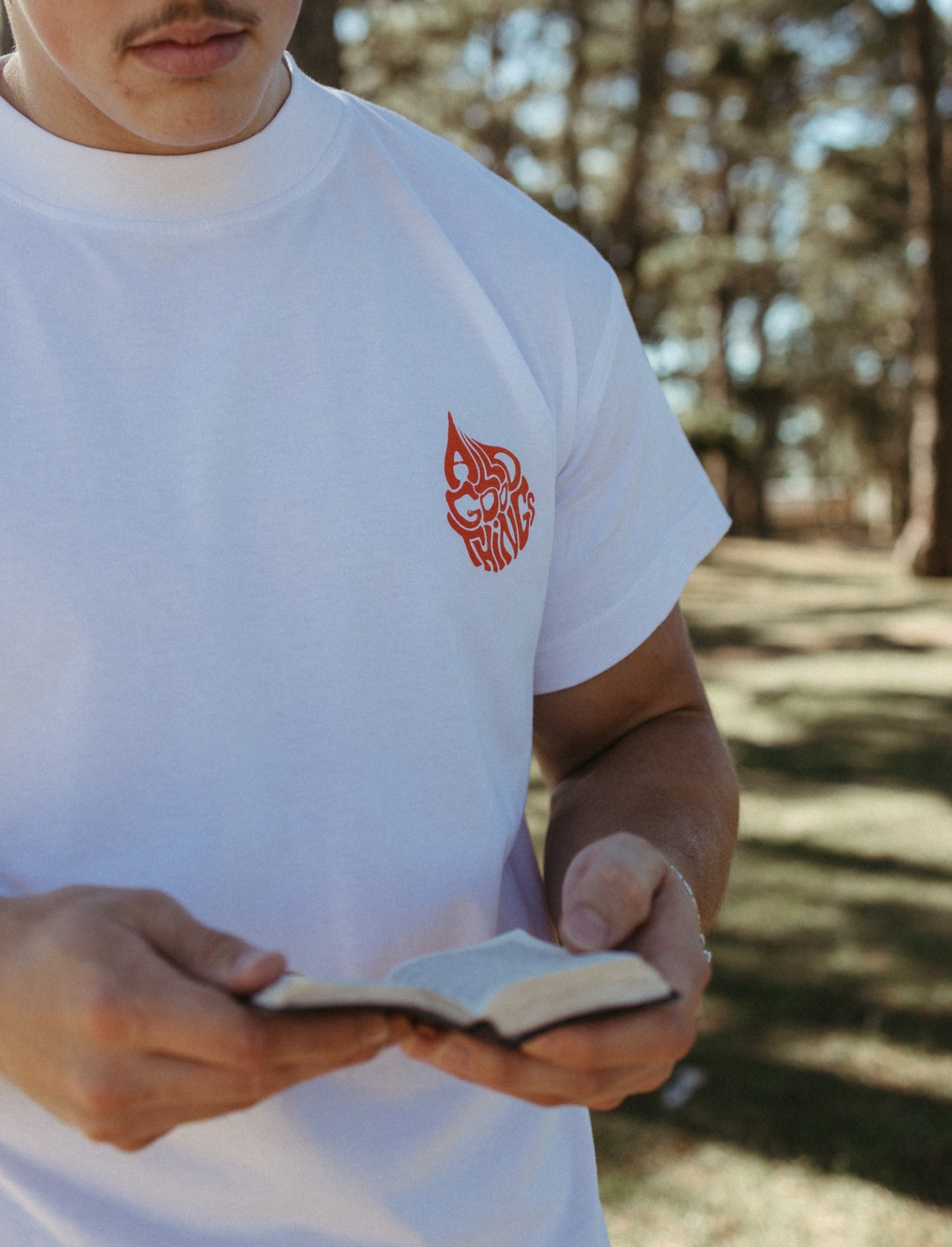 Person wearing a white Christian All Good Things t-shirt with a red logo, reading a bible outdoors.