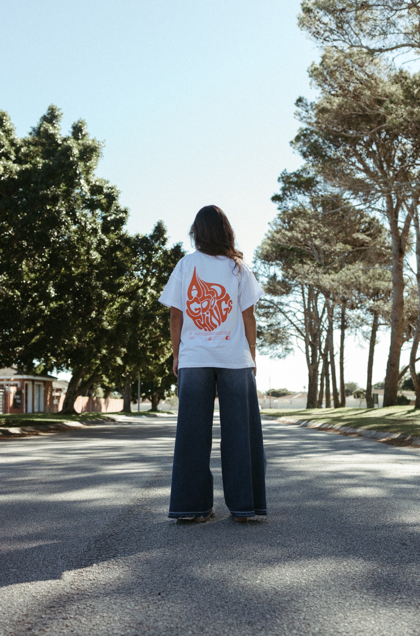 Person wearing a white All Good Things Christian t-shirt with a orange graphic design standing on a tree-lined street.